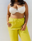 A plus-size model wearing a white lace-trimmed bralette and bright yellow-green drawstring pants, standing against a white tiled wall.