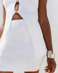 A close-up of a model wearing a white textured knit dress with cutouts and decorative gold-toned buttons down the front, paired with a chunky metallic cuff bracelet.