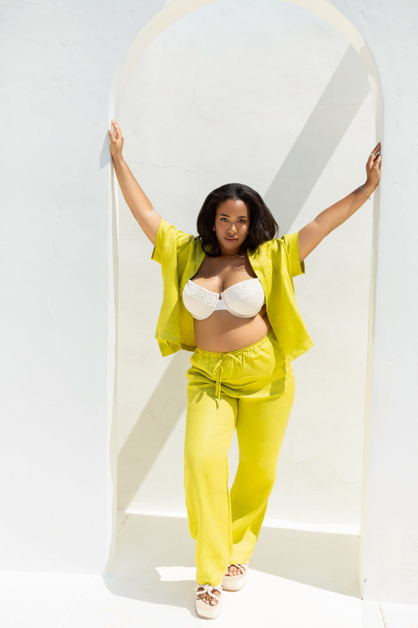 A woman posing in a bright yellow two-piece lounge set consisting of a short-sleeve top and matching drawstring pants, styled with a white bralette and white sneakers, set against a white arched wall.