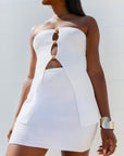 A woman wearing a white strapless mini dress with a textured knit body, structured shoulders, and a front cutout embellished with gold-tone buttons.